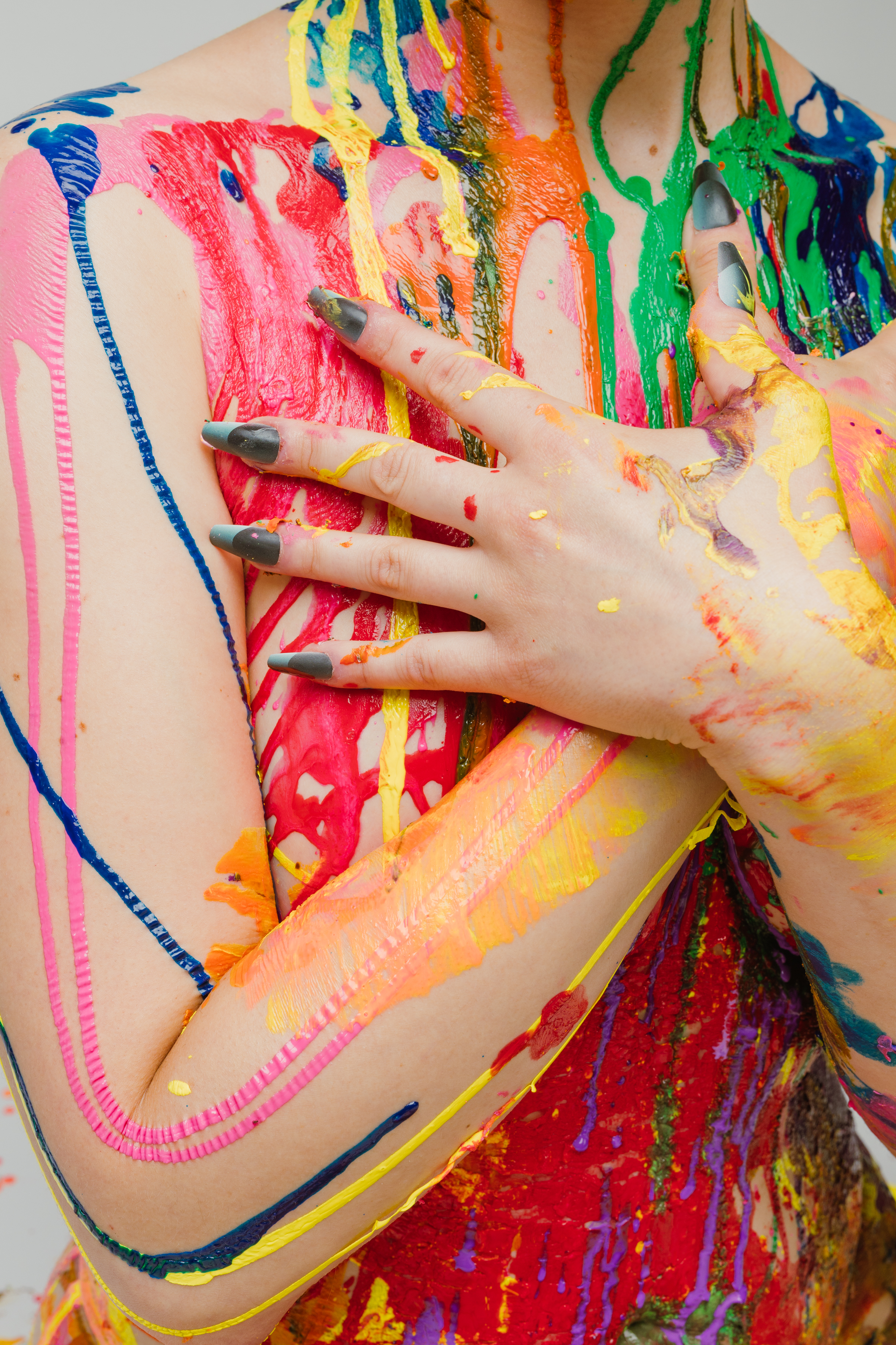 A Person with green painted nails and nail-extensions in a slightly lighter shade crosses their hands over their chest, with their skin covered in colorful paint splatters and streaks.